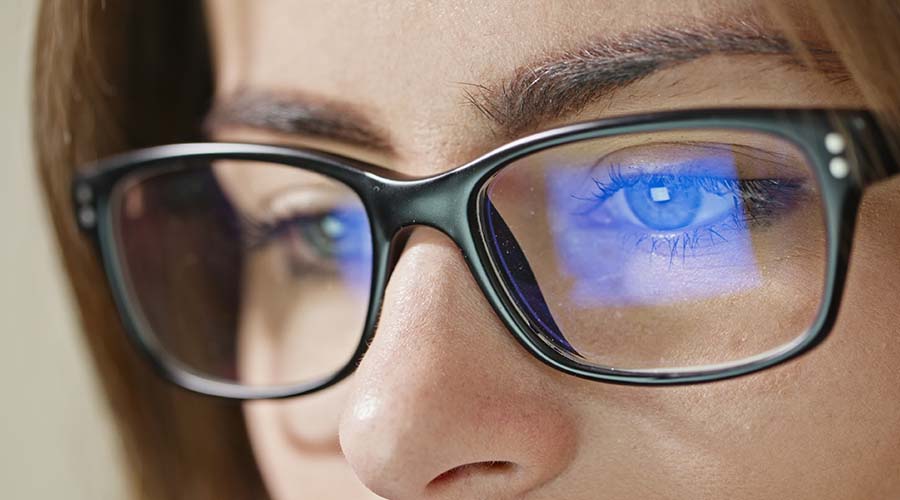 Zoom sur les yeux d'une femme portant des lunettes, reflétant la lumière bleue des écrans, soulignant l'impact de la lumière artificielle sur la peau et le contour des yeux.