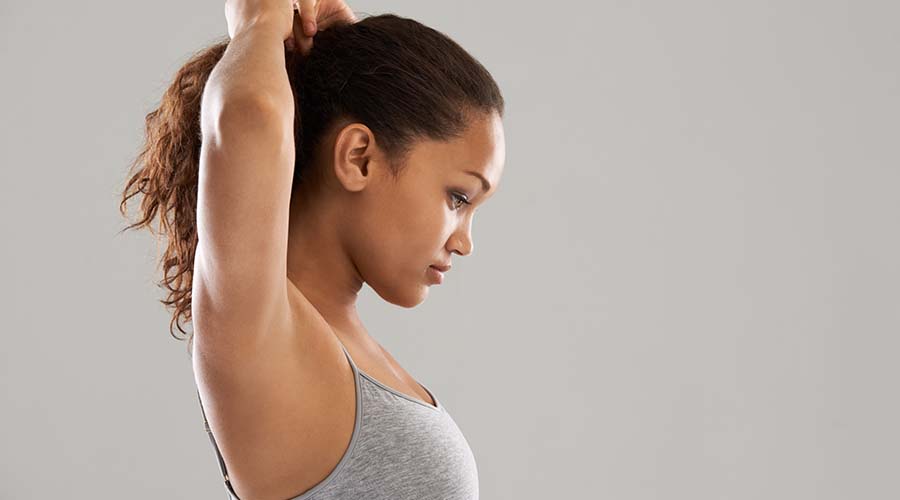 Une jeune femme s'étire avant une séance de sport, portant une tenue de fitness grise. Son visage est concentré et tourné de profil, mettant en évidence une peau lisse et éclatante.