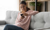 Femme pull rose sur son canapé gris qui regarde son téléphone en souriant