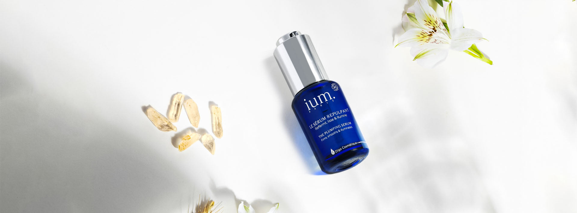 IUM Plumping Serum set against a luminous white background, surrounded by crystals and flowers, illustrating the purity, efficacy and natural firming effect of the treatment.
