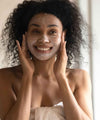 A pretty mixed-race woman smiles as she applies the ium paris mineral scrub mask in a bathroom.