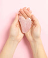 Woman's hands, seen from above, holding a rose quartz ium gua sha massage stone on a pink background